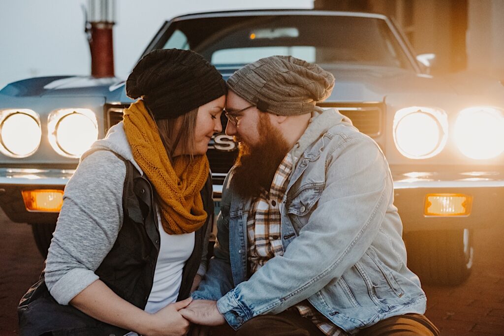 A couple in their fall clothes sit in front of their classic Chevy SS with its headlights on and touch their foreheads together while at a vintage gas station, photographed by a Chicago engagement photographer