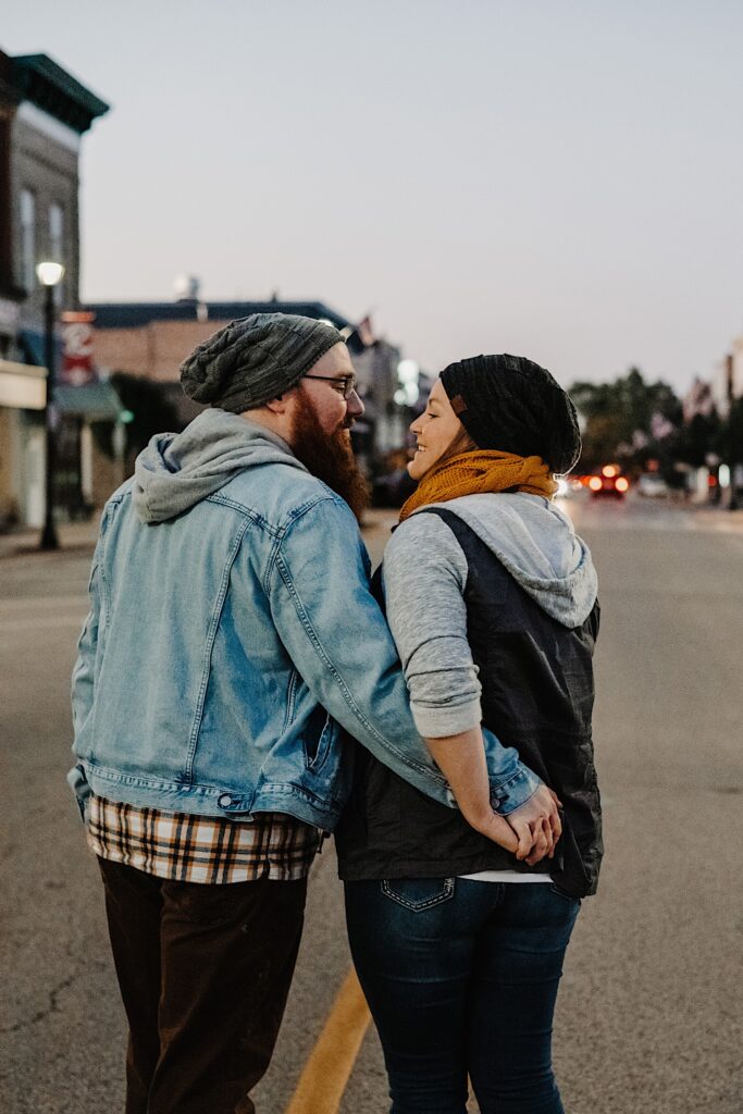 A couple in their fall clothing walk down the middle of a street in a downtown and hold hands during their engagement session