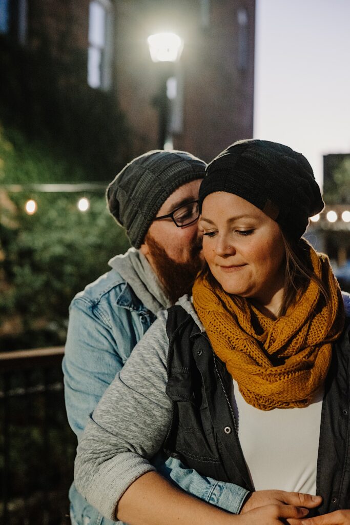 A man hugs a woman from behind and kisses her on the cheek while she smiles, they're both in their fall clothes and standing under a street light
