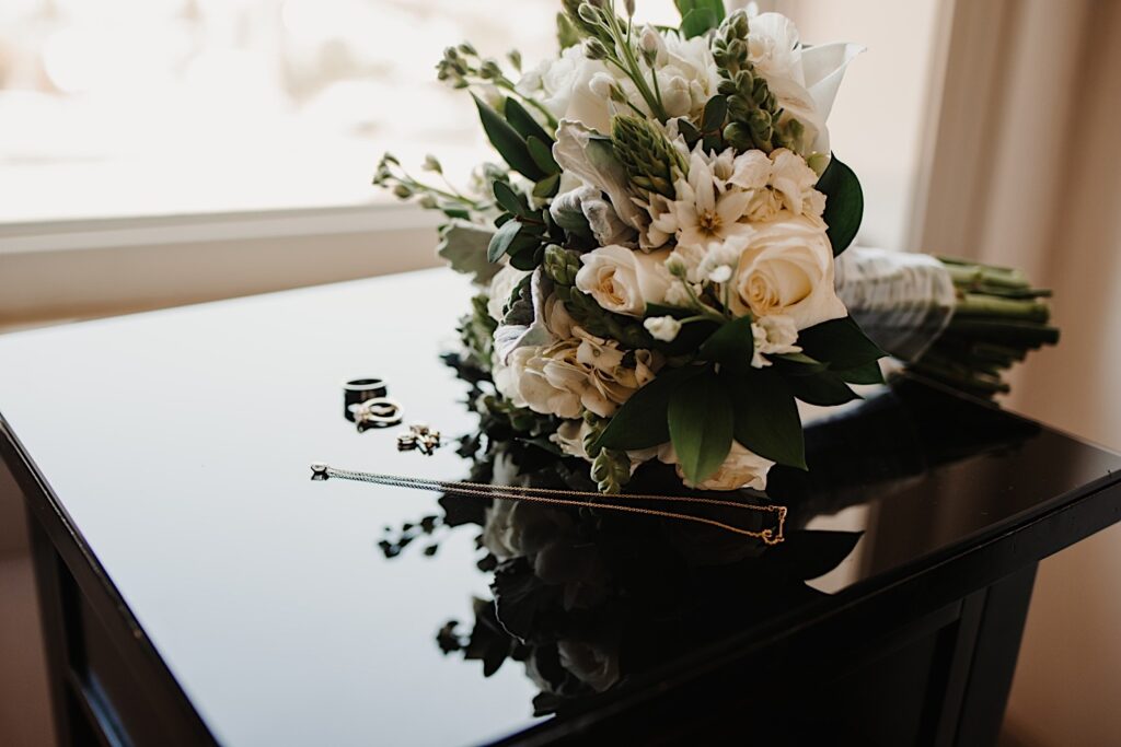 Detail photo of a necklace, wedding rings, and a bouquet laying on a black table next to a window