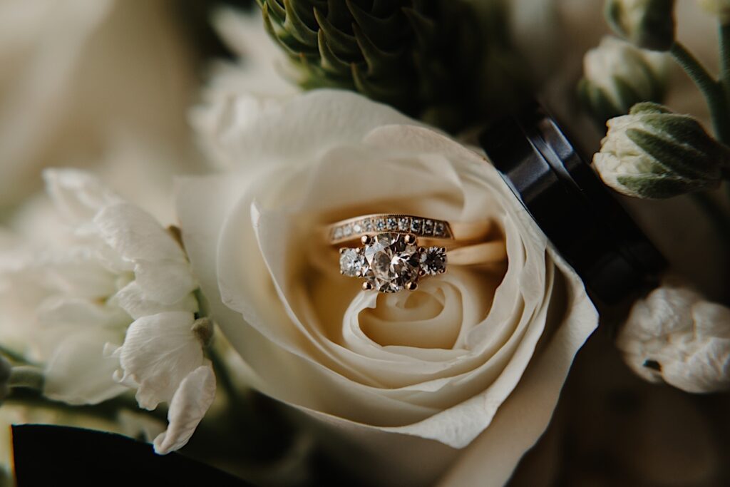 Detail photo of wedding rings resting on the inside of a white rose that is part of a bouquet