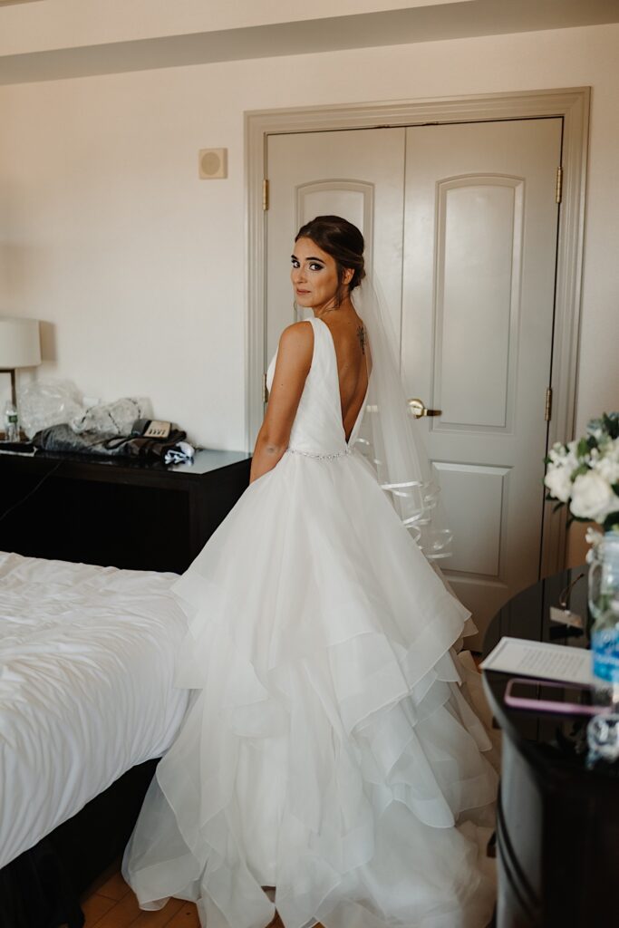 A bride facing away from the camera standing next to a bed looks back over her shoulder towards the camera