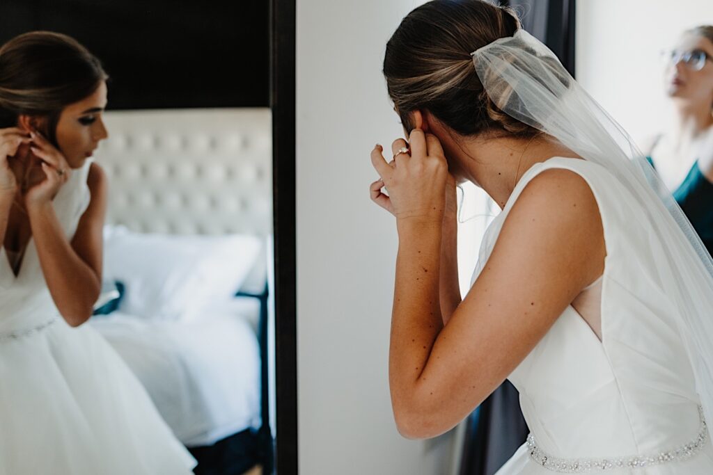 A bride facing away from the camera puts an earring on and can be seen in a mirror to her left
