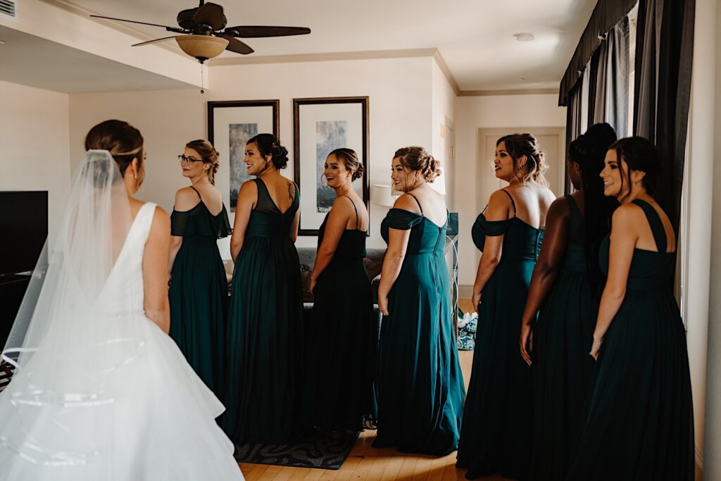 A bride faces away from the camera and stands in a room in front of her 7 bridesmaids who are all turning around to see her in her dress for the first time