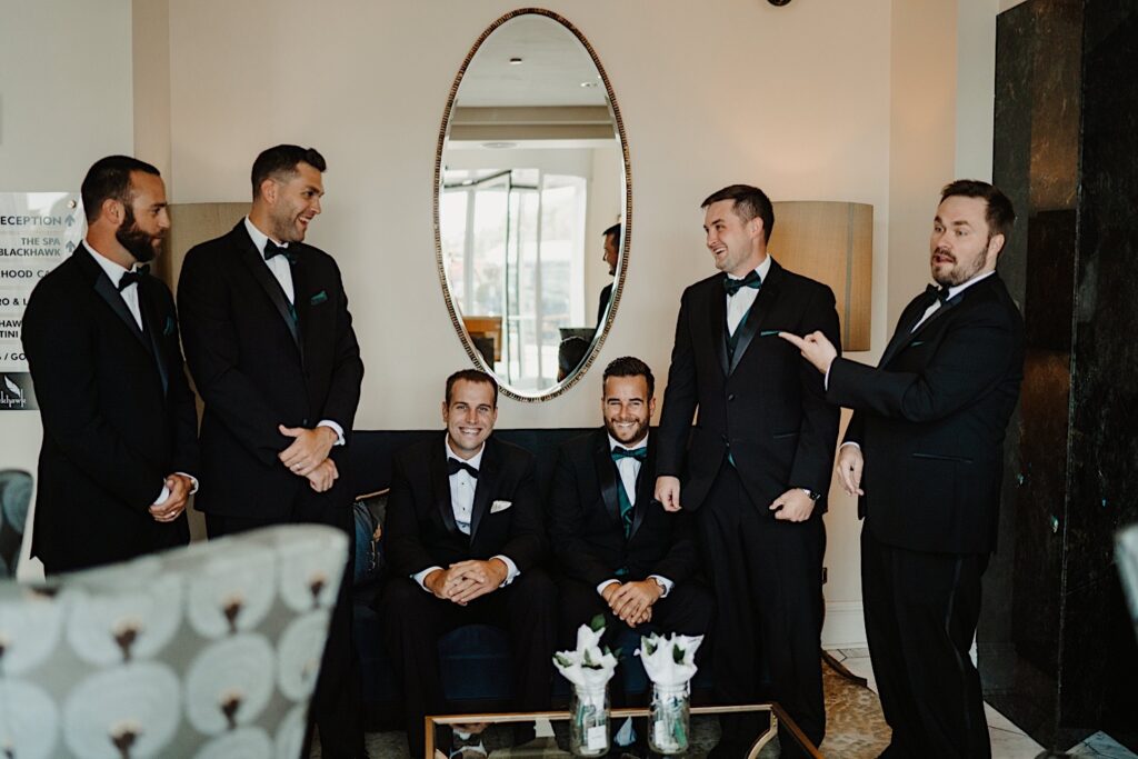 A groom sits on a black couch with a mirror over head, one of the groomsmen is sitting next to him and the other 4 groomsmen are standing on either side of the couch
