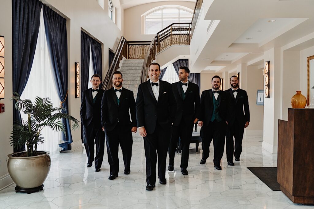 A groom walks towards the camera in a fancy hotel lobby with his 5 groomsmen walking along behind him on either side of him