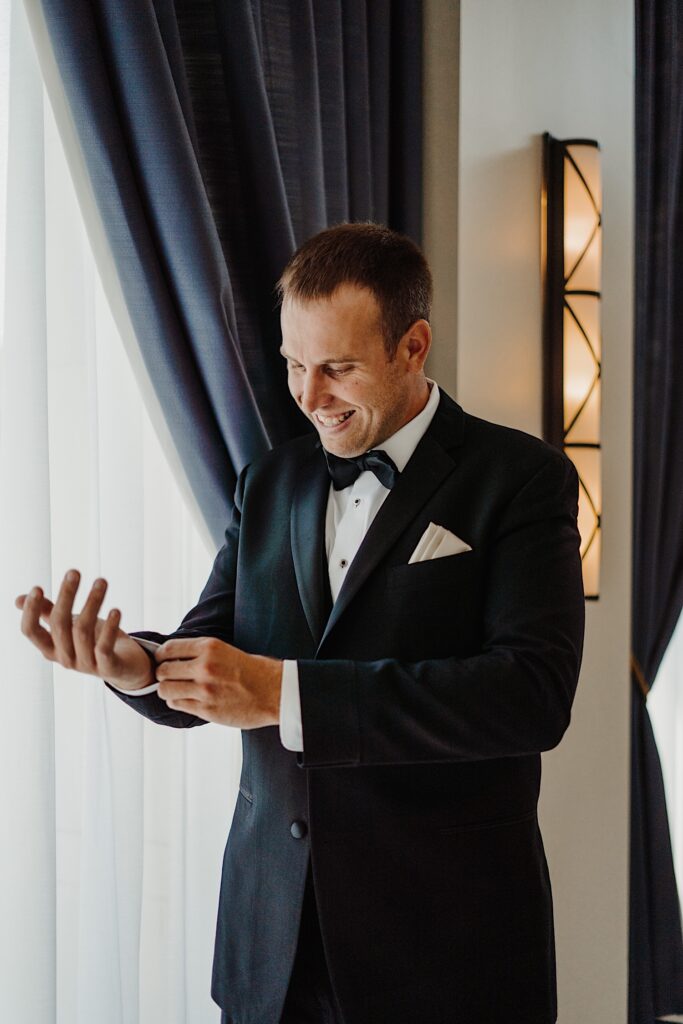 A groom smiles as he stands in front of a window and adjusts the cuff of his suit