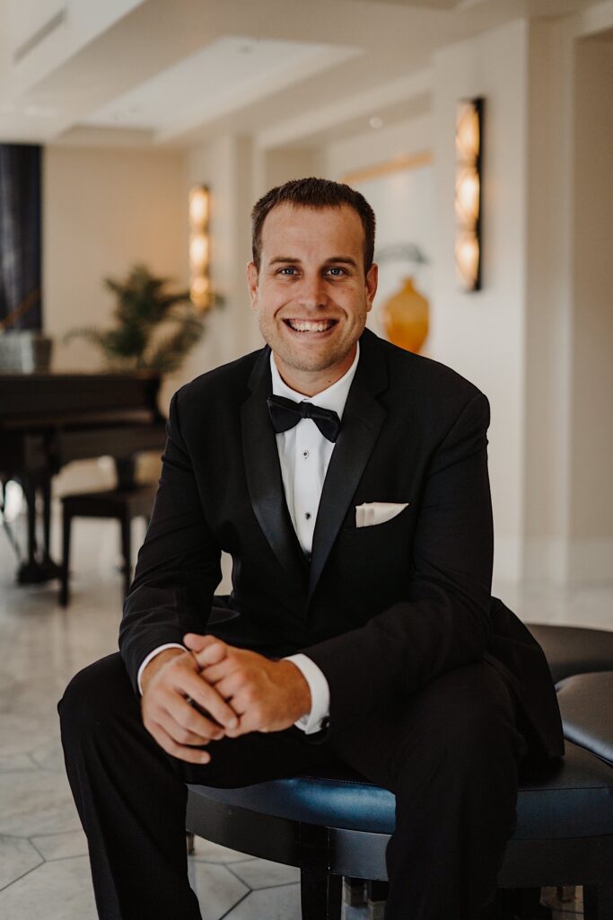 A groom sits on a bench in a fancy hotel lobby and smiles at the camera