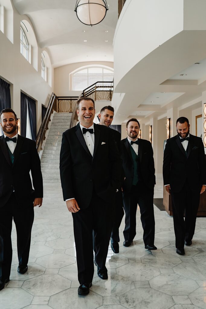 A groom smiles and laughs walking towards the camera in a fancy hotel lobby with 4 of his groomsmen walking along on either side of him