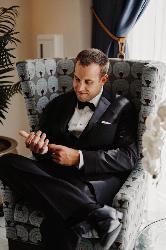 A groom sits in a chair next to a window and looks down as he adjusts the cuff of his suit