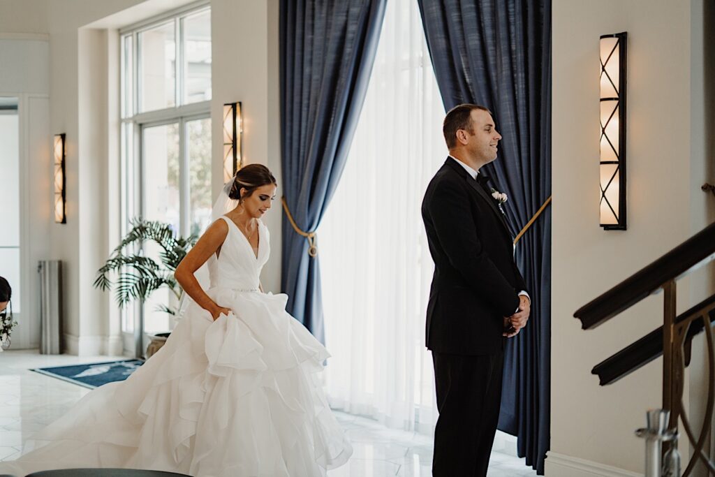 In a hotel lobby next to a large window a bride sneaks up on a groom who is smiling facing away from her as the two are about to have their first look with one another