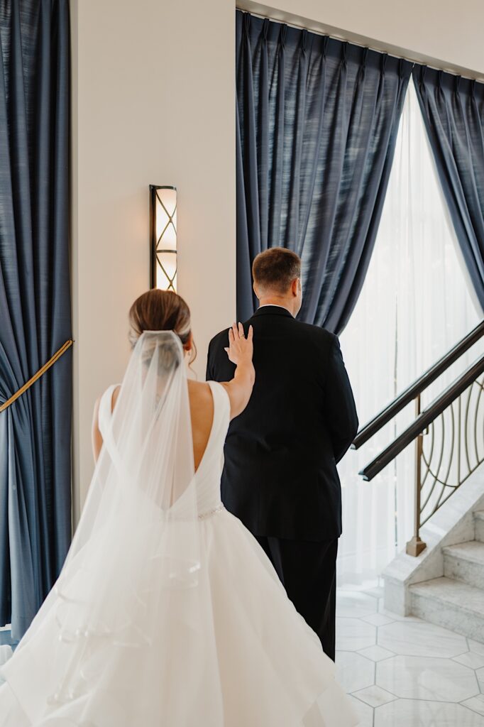 A bride and groom are facing away from the camera in a hotel lobby, the bride is tapping the groom on the shoulder for him to turn around for their first look