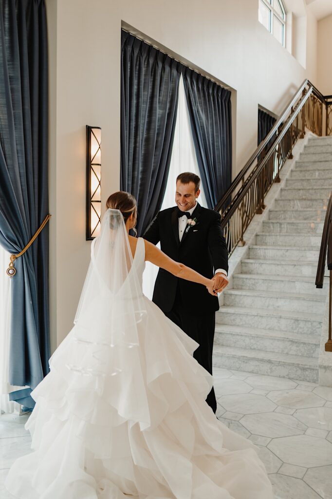 Standing in front of a large staircase a groom smiles while holding the brides hands who is facing away from the camera after the two just shared their first look