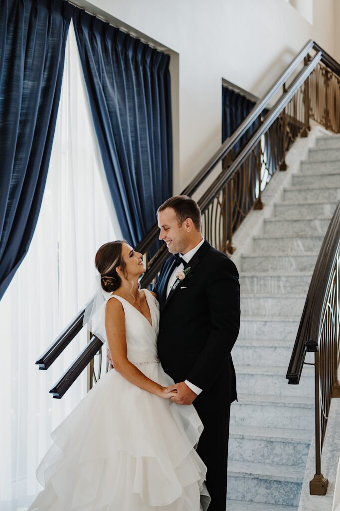 A bride and groom stand side by side and hold hands while smiling at one another in front of a staircase in a hotel lobby