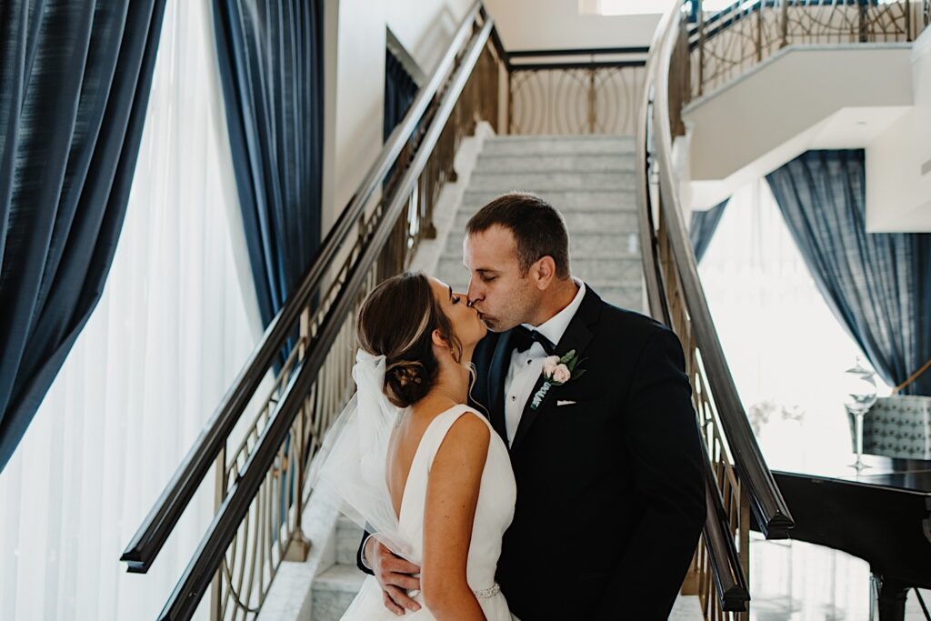 A bride and groom kiss one another while standing in front of a staircase in a fancy hotel lobby