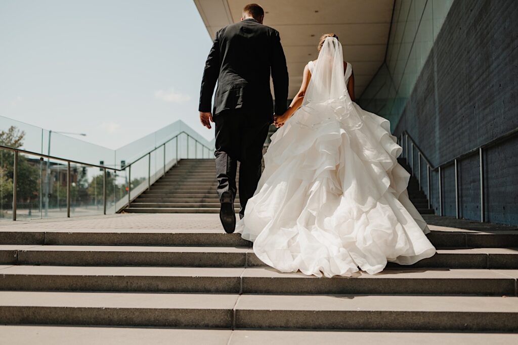 A bride and groom holding hands walk away from the camera on a large staircase outside of a building