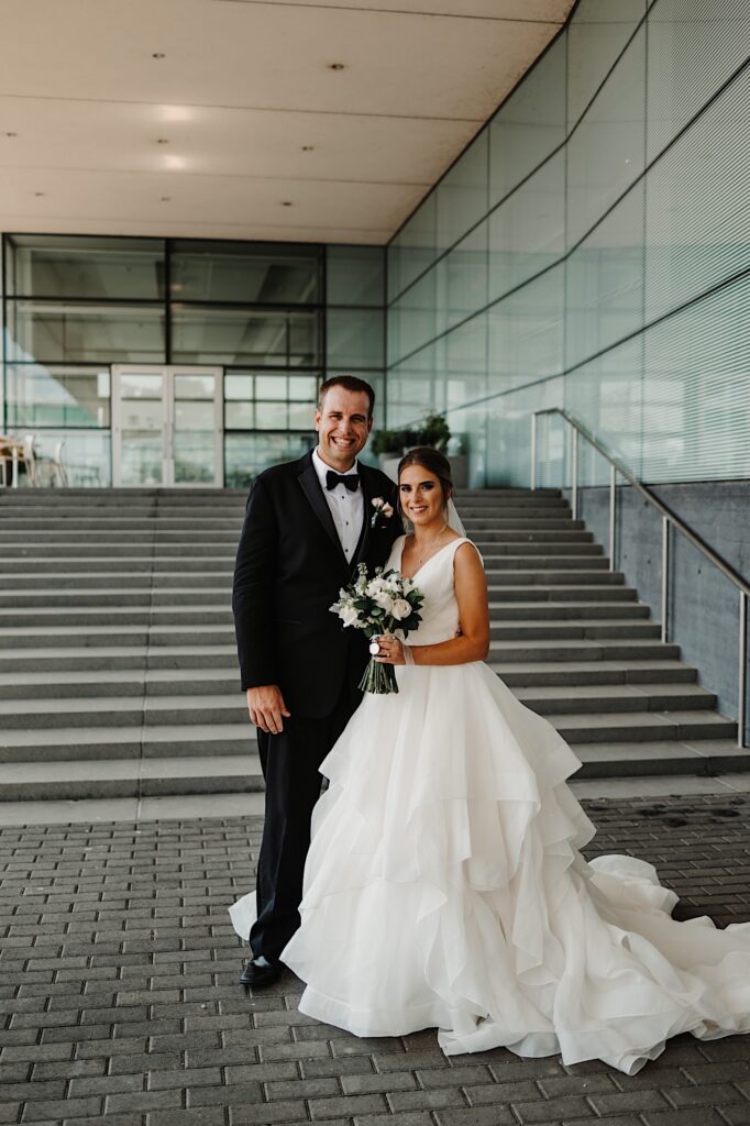 A bride and groom stand side by side for a portrait on an staircase outside of a building and smile at the camera