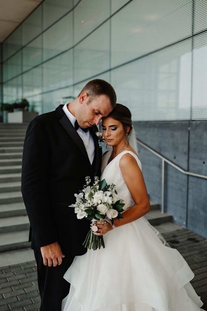A bride and groom embrace one another and look down at a flower bouquet while standing on a staircase outside of a building