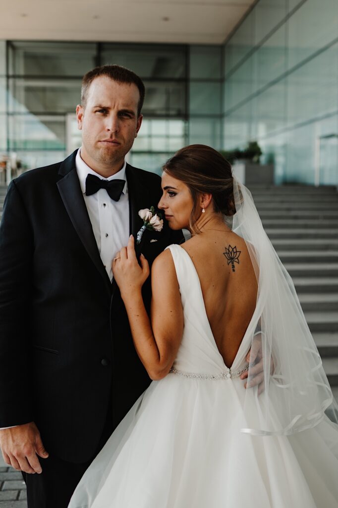 A bride facing away from the camera looks over her shoulder to smell a small bouquet, the groom is standing next to her with his hand around her waist and is looking at the camera while the two stand on an outdoor staircase