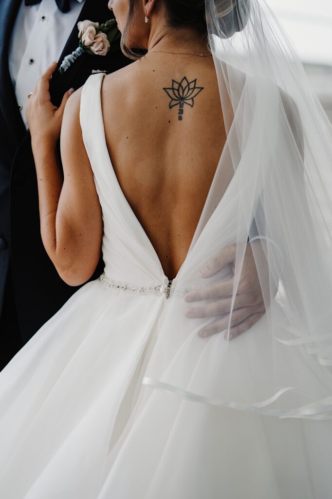 Shoulder down photo of the back of a bride in her wedding dress as she smells a small flower bouquet with the groom's hand around her waist