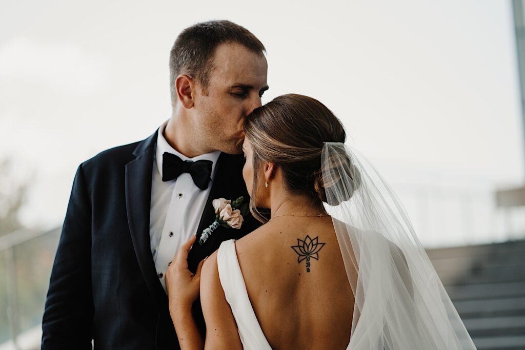 A groom kisses a bride's forehead as she has her back to the camera while the two stand on an outdoor staircase