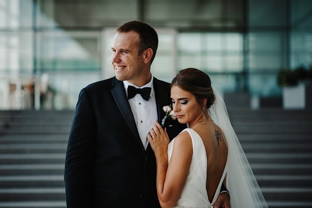 A bride faces away from the camera towards the groom and looks down over her shoulder as the groom looks to the left and smiles while the two stand on an outdoor staircase