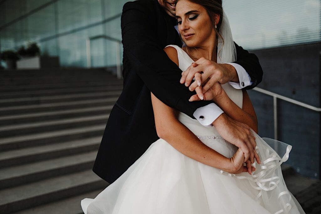 A bride looks back over her shoulder as a groom smiles while hugging her from behind as the two stand on a staircase outdoors