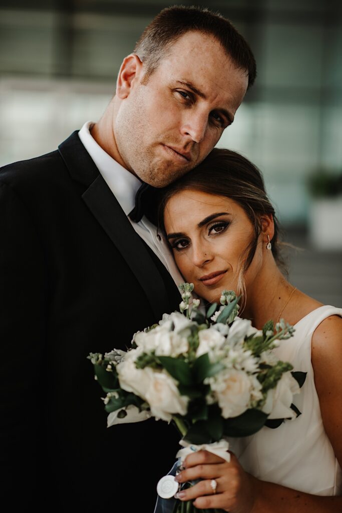 Portrait photo of a bride resting her head on the grooms chest and holding up her bouquet while they both look at the camera