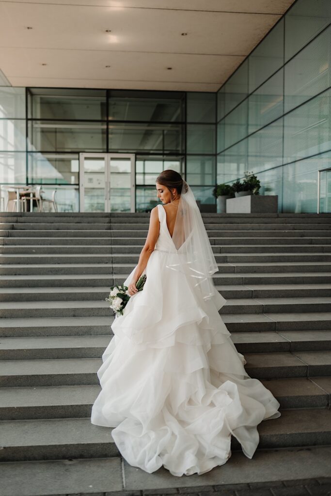 A bride facing away from the camera looks down at her bouquet while walking up some stairs outside a building