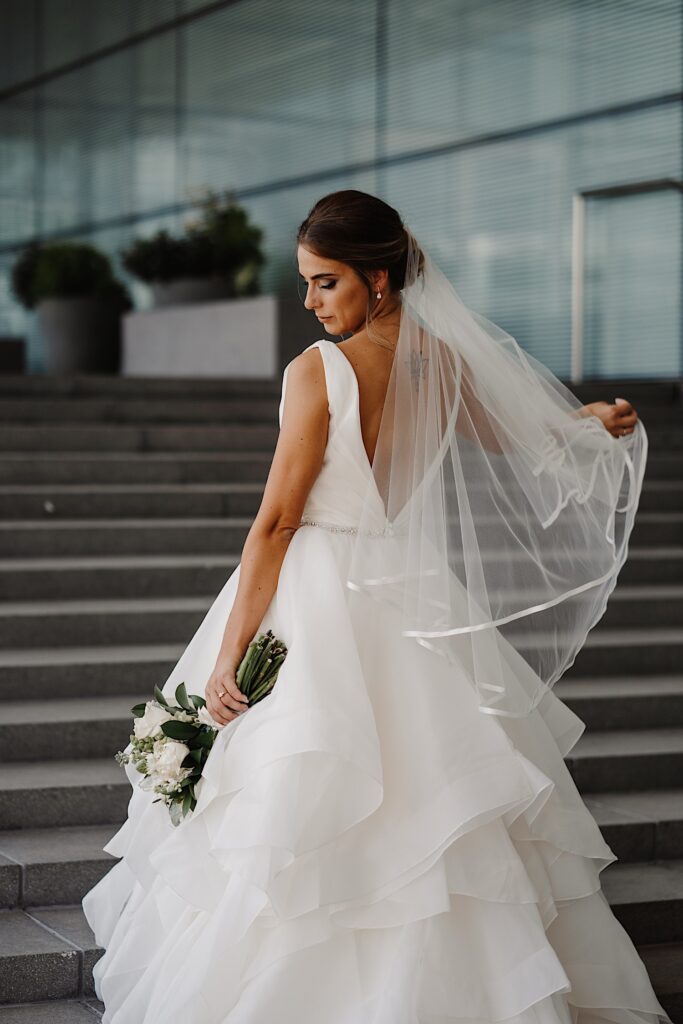 A bride facing away from the camera on a set of stairs looks down at her bouquet and plays with her veil in her other hand