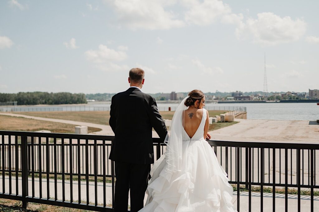 A bride and groom facing away from the camera lean on a railing and look out over a body of water out in front of them