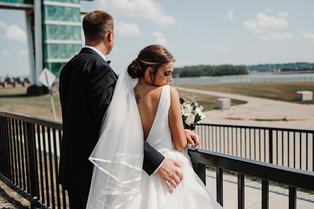 A bride and groom face away from the camera and lean on a railing, the groom has his arm around the bride and is looking out over the view while the bride looks over her shoulder at the grooms hand on her