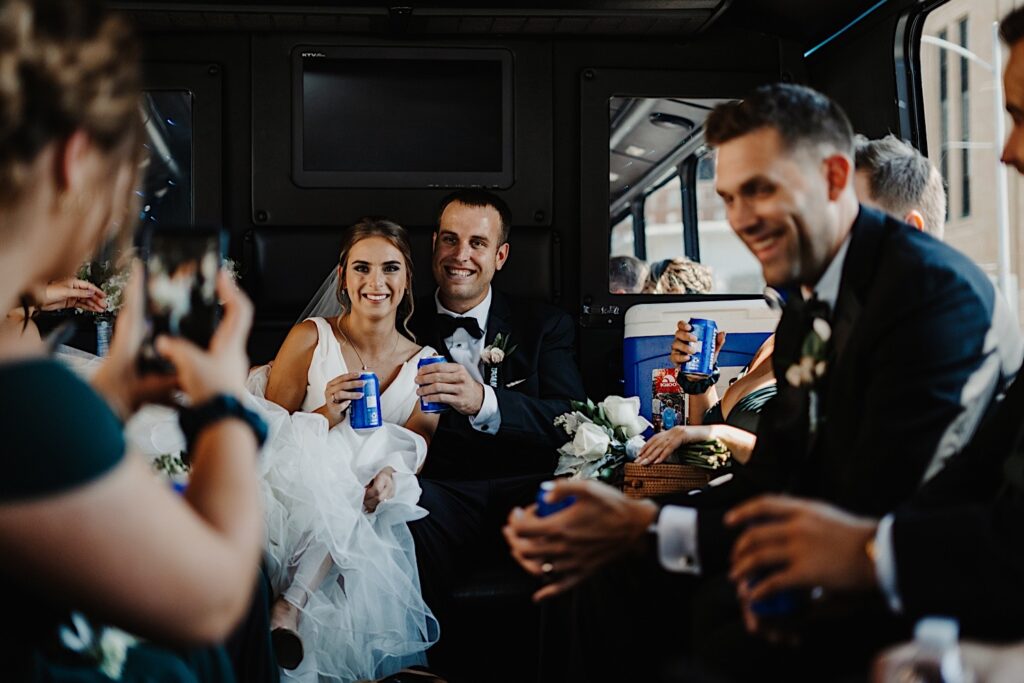 A bride and groom sit next to one another and smile while on a party bus with their wedding parties, the wedding party members are laughing and everyone has a bud light in their hand