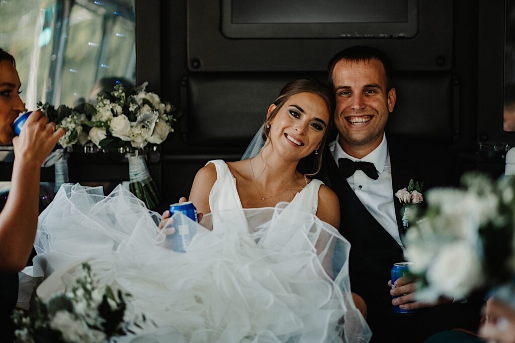 A bride and groom sitting next to one another on a party bus smile at the camera with Bud lights in their hand