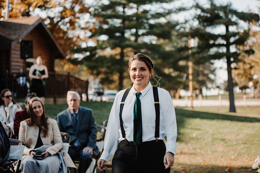 A woman in a shirt and tie smiles while walking down the aisle before an intimate wedding ceremony
