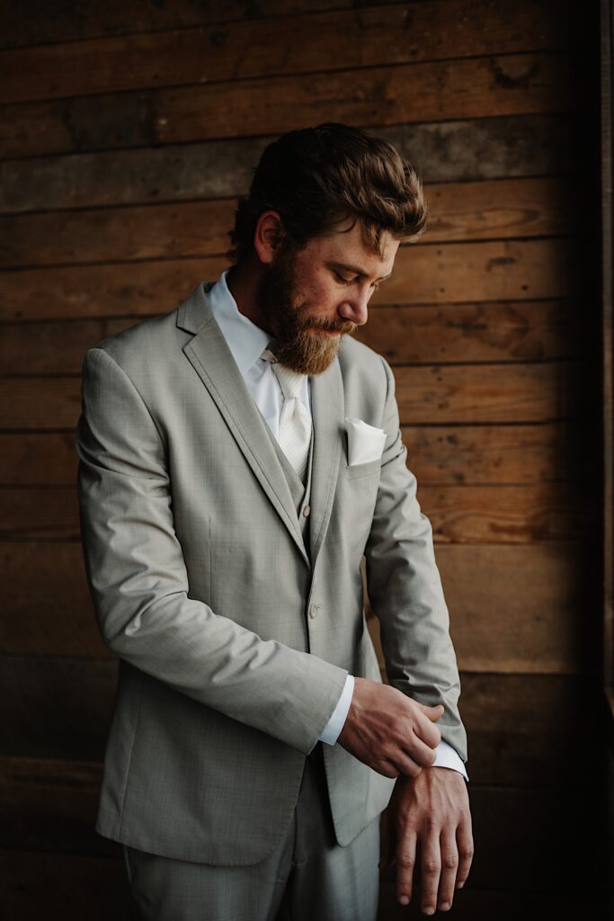A groom looks down and adjusts the cuff of his suit coat while standing in front of a wood panel wall