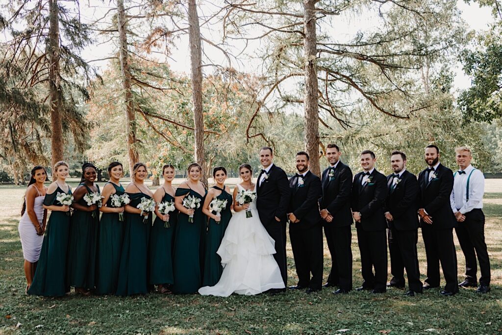 A bride and groom stand with their wedding parties on either side of them as everyone smiles at the camera in front of a line of trees