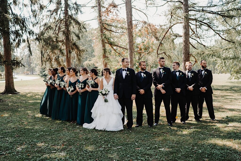 A bride and groom stand next to one another and hold hands while looking in opposite directions, their wedding parties are on their respective sides and the group is standing in front of a line of tall trees
