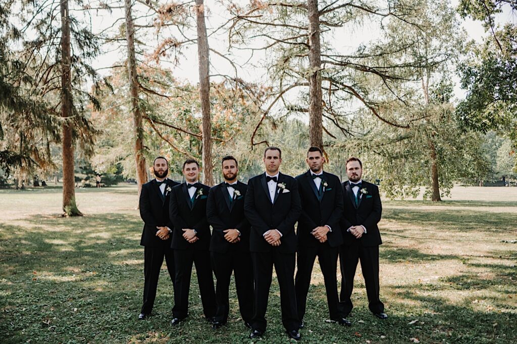 A groom stands with his hands clasped over one another with his 5 groomsmen in the same pose on either side of him all looking at the camera in front of a line of trees