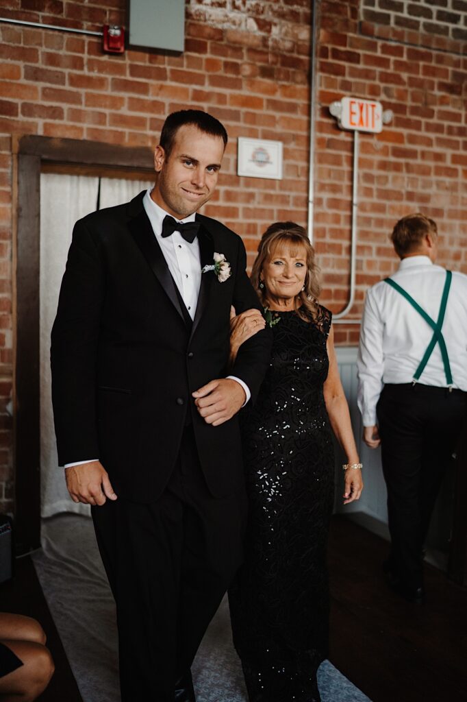 A groom walks into the room of his wedding ceremony with his mom walking alongside him