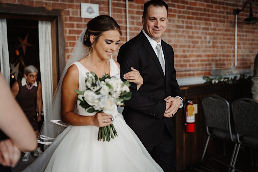 A bride enters the indoor ceremony space at Stardust in Davenport during her wedding ceremony, she smiles as she is walked into the room by her father