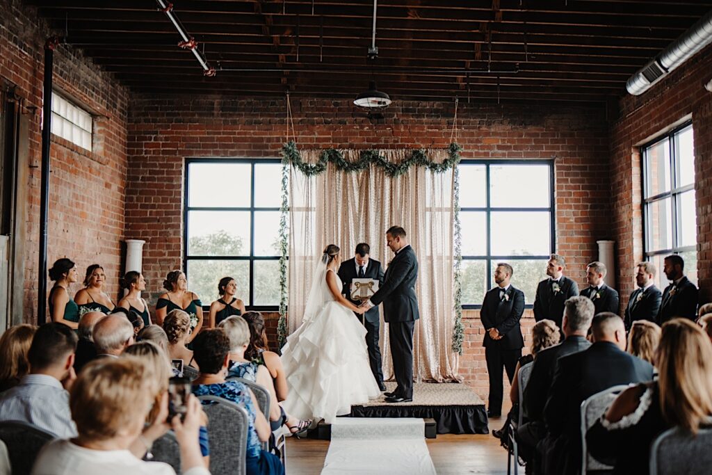 At a wedding ceremony at Stardust in Davenport a bride and groom hold hands while the officiant talks and their guests are seated watching