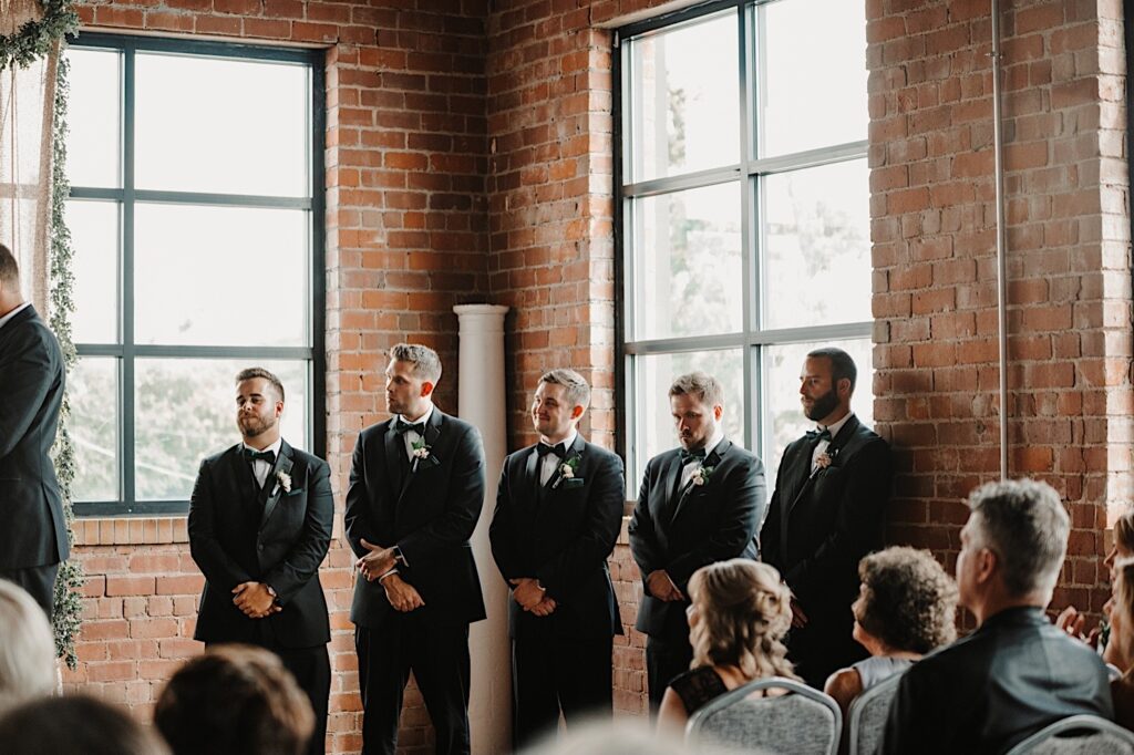 5 groomsmen stand to the side and have their hands clasped watching a wedding ceremony take place at Stardust in Davenport