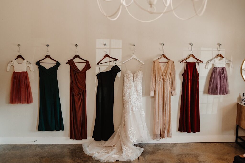 A wedding dress and 7 other dresses hang on a wall in a getting ready space before a wedding, photographed by a Central Illinois wedding photographer
