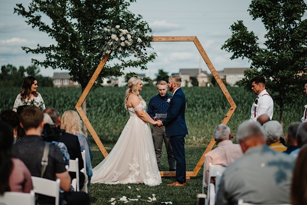 Guests and wedding parties watch as a bride and groom hold hands during their outdoor wedding ceremony at their wedding venue, Destihl Brewery in Normal, Illinois