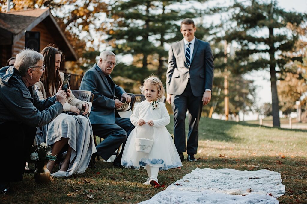 A flower girl walks down the aisle of an intimate wedding ceremony in the fall next to a cabin near Bloomington Illinois as guests watch and take photos of her
