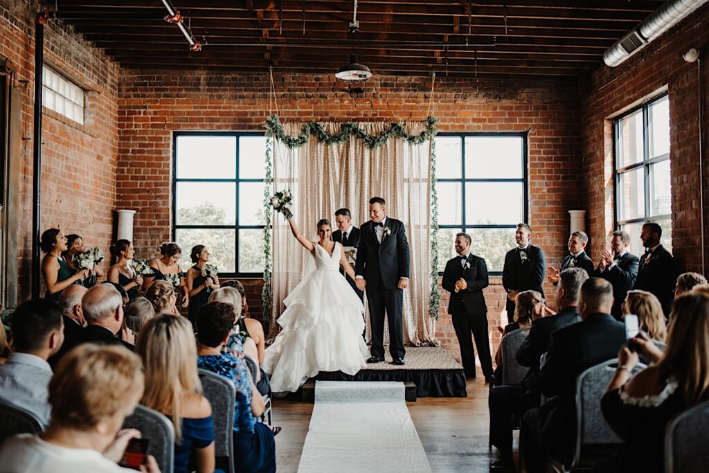 A bride lifts her bouquet in the air and smiles while holding the grooms hand as the two begin to exit their wedding ceremony while their guests and wedding parties cheer for them