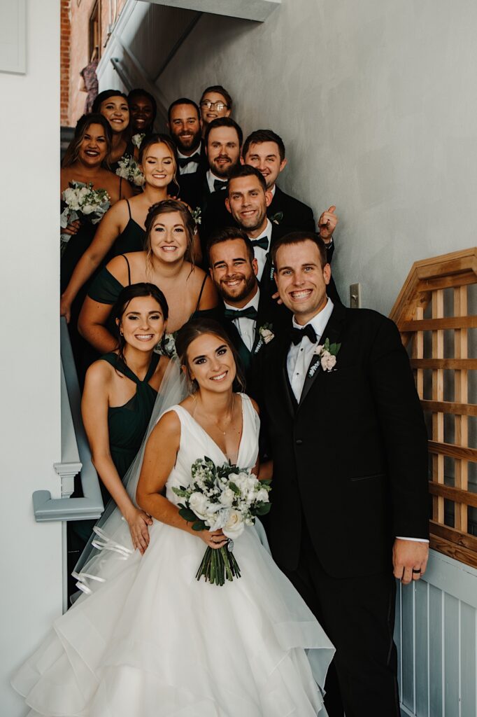 A bride and groom stand next to one another and smile at the camera, behind them are their wedding party members standing on a staircase all smiling at the camera