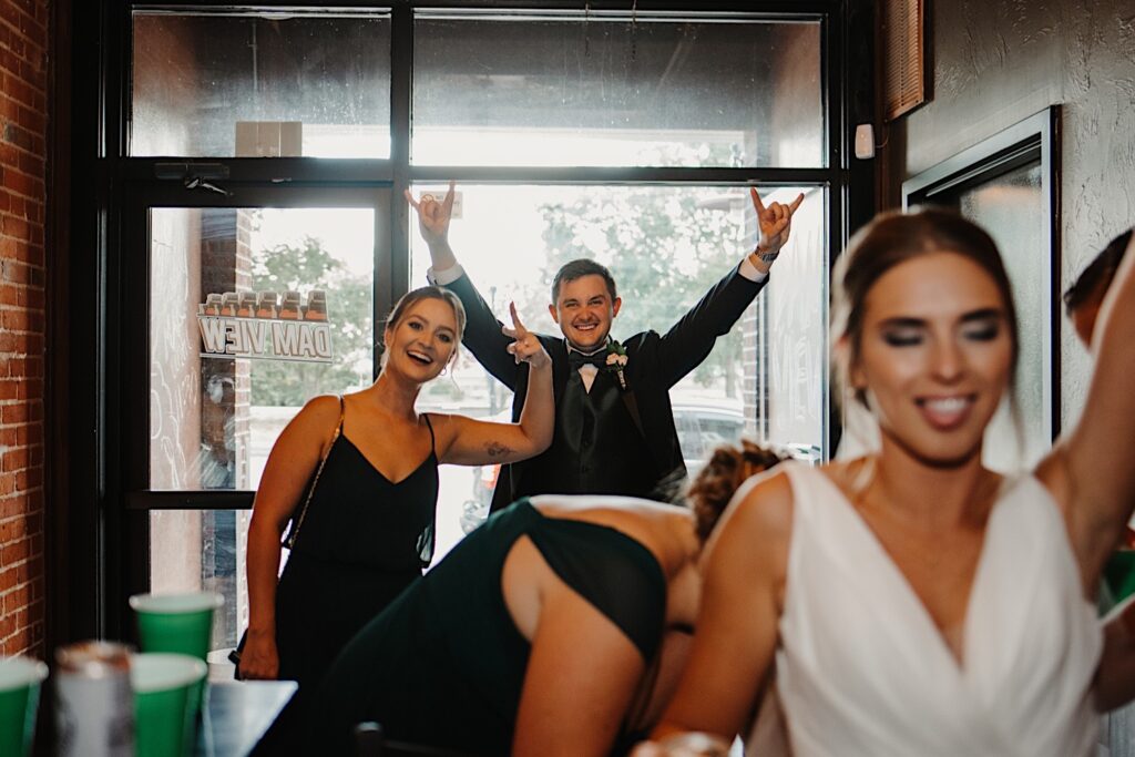 A bride smiles and lifts her hand in the air while a groomsmen and bridesmaid in the background smile and gesture at the camera