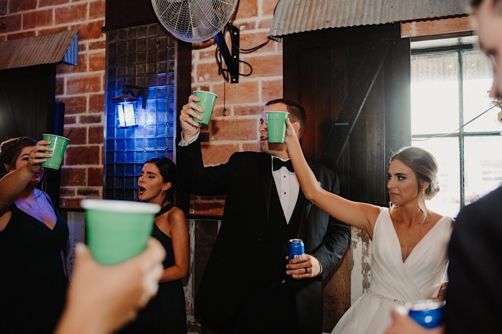 A bride and groom raise green solo cups in the air in a brick room at Stardust in Davenport with their wedding party members around them all doing the same